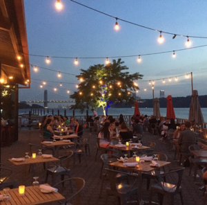 The Hudson's outside deck at twilight, as strands of lights crisscross above people attending a milestone event. it is a beautiful evening