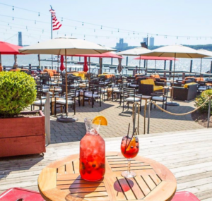 an enticing red drink in a sleek pitcher sits on a circular table in the foreground, tables and chairs under sun umbrellas sit in the background on a gorgeous summer day. it is on the outside deck of The Hudson
