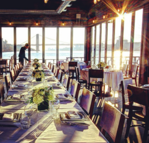 The interior of The Hudson's large event hall, set up for a grand gathering. There are long tables with white tablecloths, with chairs on each side. You can see the Hudson River and George Washington Bridge in the background through floor-to-ceiling windows.